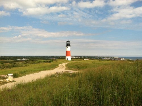 A lighthouse stands on a grassy path, with a blue sky and distant coastline in the background.