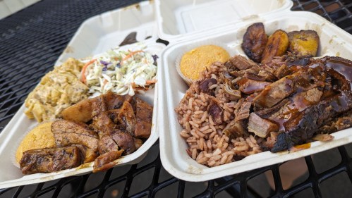 Two takeout containers filled with rice, beans, grilled meats, plantains, coleslaw, and cornbread on a black table.