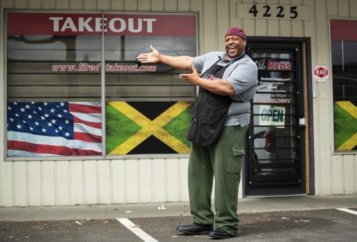A smiling man in an apron stands outside a takeout restaurant with Jamaican and American flags in the window.