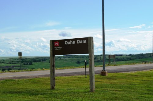 Sign for Oahe Dam with green fields and blue sky in the background, near a road.