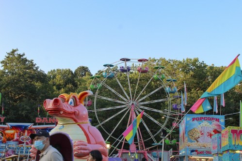 A colorful carnival scene featuring a pink pig ride, a Ferris wheel, and festive flags under a clear blue sky.