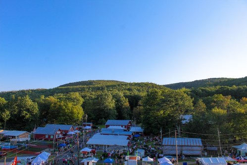 Aerial view of a bustling outdoor market surrounded by trees and hills under a clear blue sky.