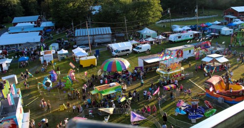 Aerial view of a vibrant fairground with colorful tents, rides, and crowds enjoying various attractions and activities.