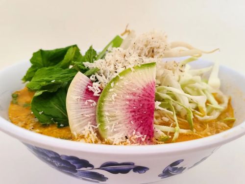 A bowl of vibrant ramen topped with mint, cabbage, radish, and shredded coconut in a flavorful broth.
