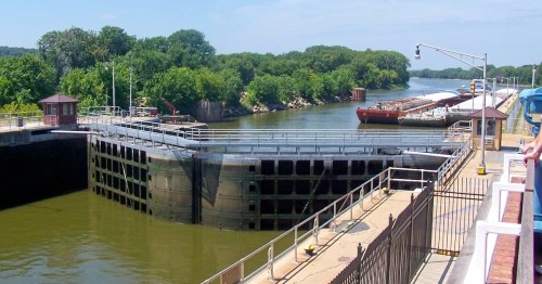 View from the observation deck of a massive river lock gate with cargo barges lined up in the waterway.
