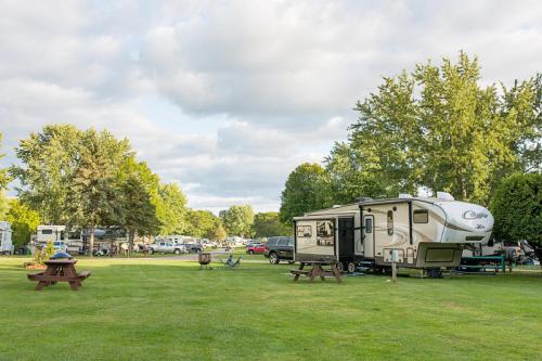 A spacious RV park with green grass, picnic tables, and trees under a partly cloudy sky. RVs are parked nearby.