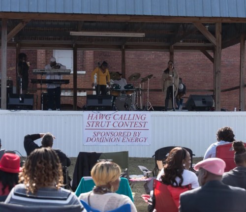 A stage with musicians performing at a strut contest, with an audience seated in front, under a blue roof.