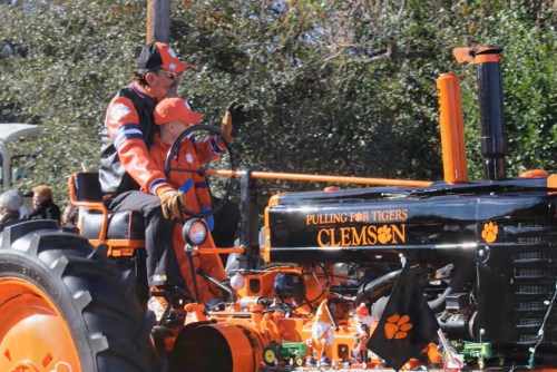 Two people in orange and black attire ride a vintage tractor decorated with "Clemson" and a tiger paw flag.