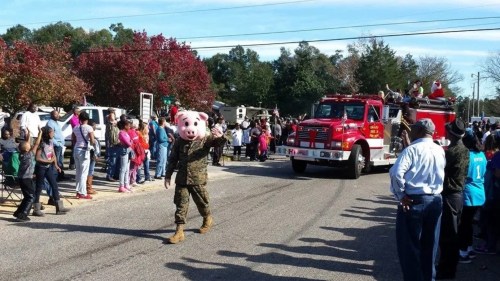 A person in a pig mask walks in a parade alongside a fire truck, with a crowd watching on both sides of the street.