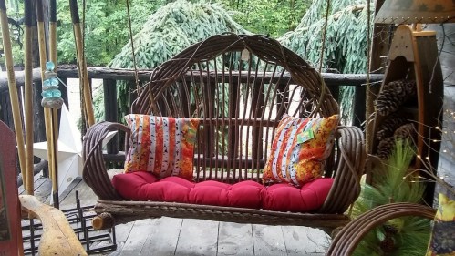 A cozy swing made of woven branches, featuring colorful pillows and a red cushion, set in a lush outdoor environment.