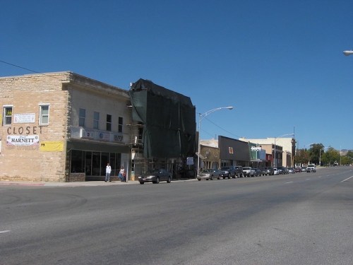 A street view of a small town with buildings, some under renovation, and parked cars along the road on a clear day.