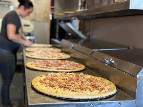 A pizza kitchen with several uncooked pizzas topped with ingredients, and a person preparing food in the background.
