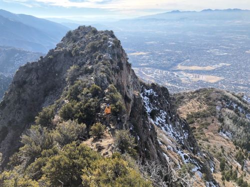A hiker navigates a rocky mountain ridge with a valley and distant mountains in the background. Snow is visible on the ground.