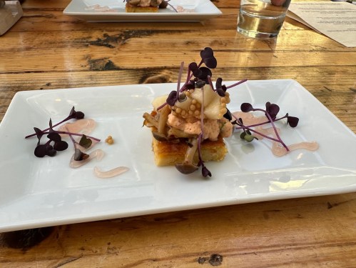 A gourmet dish featuring layered ingredients on a white plate, garnished with microgreens and sauces, set on a wooden table.