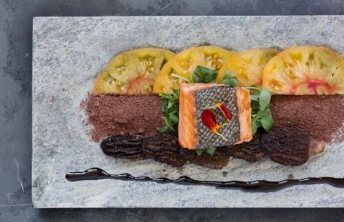 A gourmet dish featuring salmon, yellow tomatoes, greens, morel mushrooms, and a chocolate sauce on a stone plate.