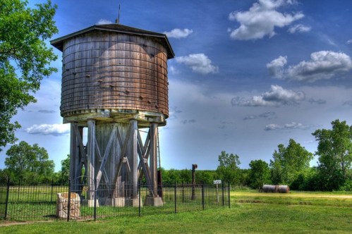 A weathered wooden water tower stands on stilts, surrounded by green trees and a blue sky with fluffy clouds.