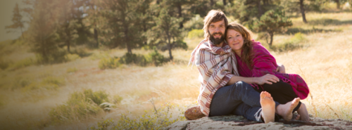 A couple sits together on a rock in a sunny, grassy field, wrapped in blankets and smiling at the camera.