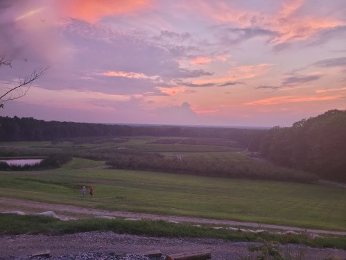 A serene landscape at sunset, featuring rolling fields, a pond, and two people walking along a path.