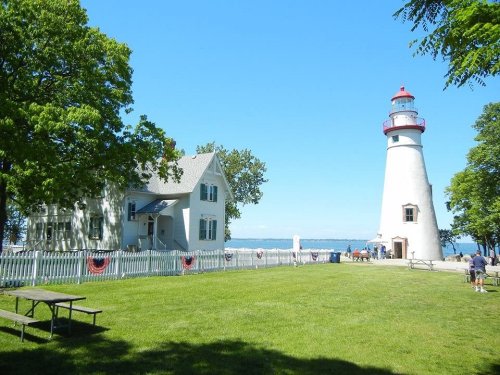 A white lighthouse beside a historic house, surrounded by green grass and trees, with a clear blue sky in the background.