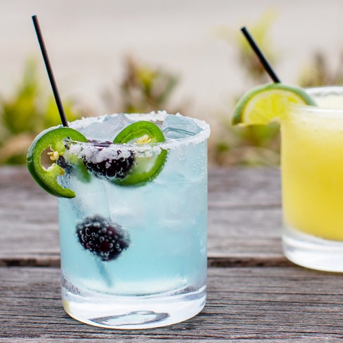 A refreshing blue cocktail with blackberries and lime, garnished with jalapeño slices, on a wooden table.