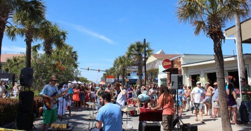 A lively street festival with a crowd enjoying live music under palm trees and a clear blue sky.