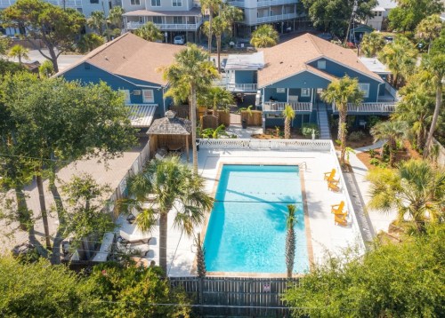 Aerial view of a pool surrounded by palm trees and colorful houses, with lounge chairs on the deck.