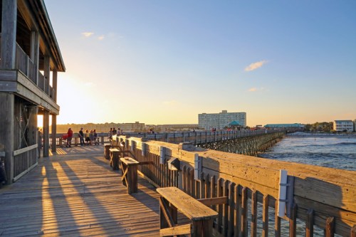 A wooden pier extends into the water at sunset, with people enjoying the view and buildings in the background.