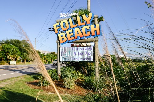 Welcome sign for Folly Beach with digital display, surrounded by greenery and palm trees on a sunny day.