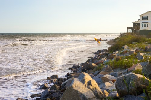A beach scene with waves, rocky shore, and two surfers walking towards the water near a house.