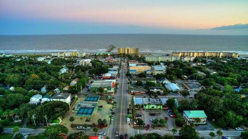 Aerial view of a coastal town with a street lined by buildings, trees, and a pier extending into the ocean at sunset.
