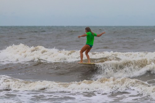 A surfer in a green shirt rides a wave on a surfboard, with ocean waves crashing around her.