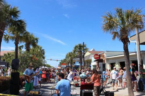 A lively street scene with a crowd enjoying live music under palm trees on a sunny day.