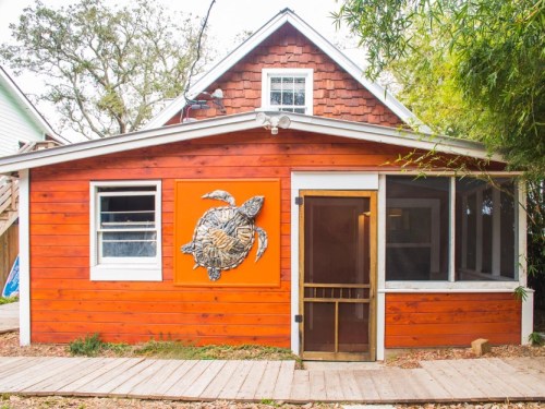 A charming orange wooden house with a turtle sculpture on the wall and a screened porch. Surrounded by greenery.