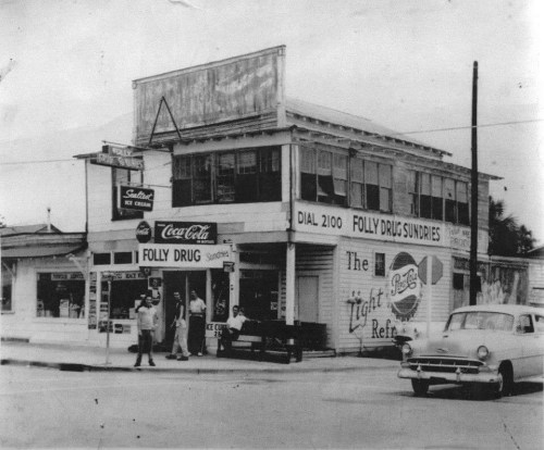 Black and white photo of a vintage drugstore with a Coca-Cola sign and a classic car parked outside.