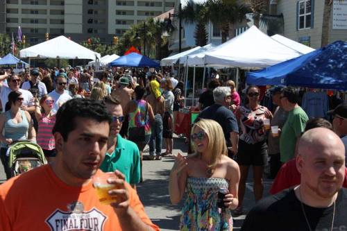 A lively outdoor festival scene with crowds, tents, and people enjoying food and drinks under sunny weather.