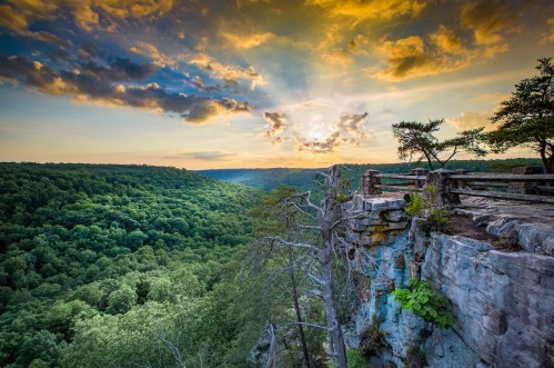 A stunning sunset over a lush green valley, viewed from a rocky cliff with dramatic clouds in the sky.