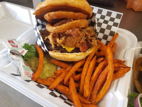 A hearty burger topped with pulled pork and onion rings, served with sweet potato fries and condiments in a takeout container.