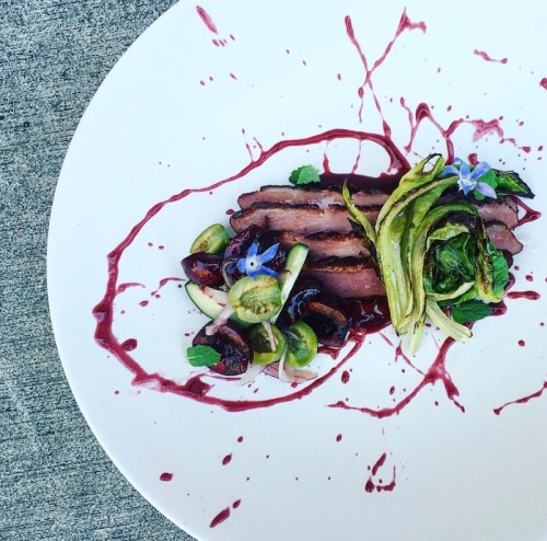 A gourmet dish featuring sliced meat, garnished with greens, fruits, and colorful sauces on a white plate.