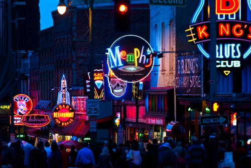 Vibrant neon signs illuminate a bustling street in Memphis at night, showcasing bars and music venues.