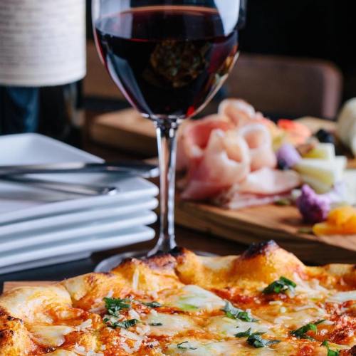 A close-up of a pizza topped with herbs, alongside a glass of red wine and a charcuterie board in the background.