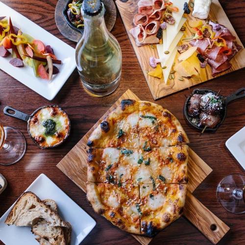 A wooden table spread with pizza, charcuterie, cheese, pickled vegetables, and drinks.