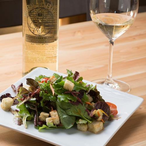 A fresh salad with greens, croutons, and tomatoes, accompanied by a glass of white wine and a bottle in the background.