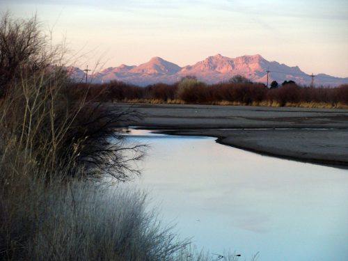 A serene landscape featuring a calm river reflecting mountains in the background, surrounded by sparse vegetation.