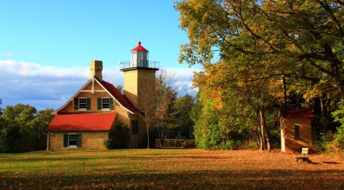A historic lighthouse beside a stone house, surrounded by colorful autumn trees and a clear blue sky.