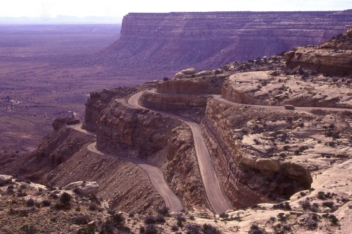 Winding road along a rocky cliff with expansive desert landscape and distant mesas under a hazy sky.