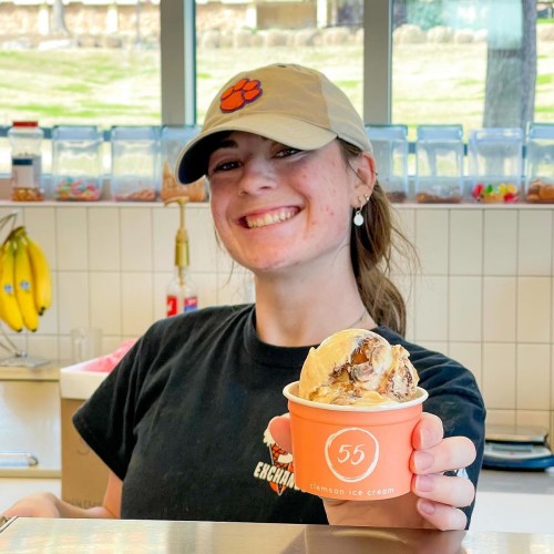 A smiling young woman in a cap holds a cup of ice cream at an ice cream shop.