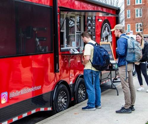 Two people with backpacks order food at a red food truck, with a woman waiting nearby.