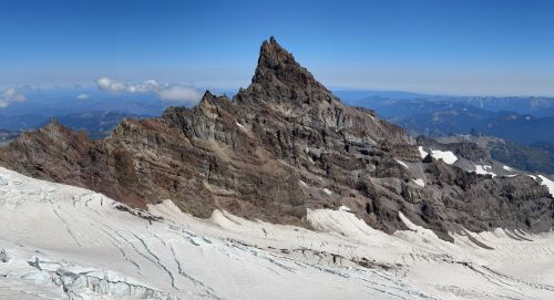 A rugged mountain peak rises above a snowy landscape under a clear blue sky, with distant mountains in the background.