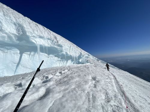 A climber traverses a snowy mountain ridge with a steep ice wall under a clear blue sky.