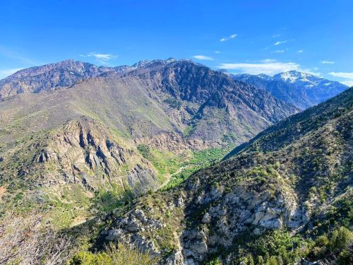 A panoramic view of rugged mountains with green valleys and snow-capped peaks under a clear blue sky.